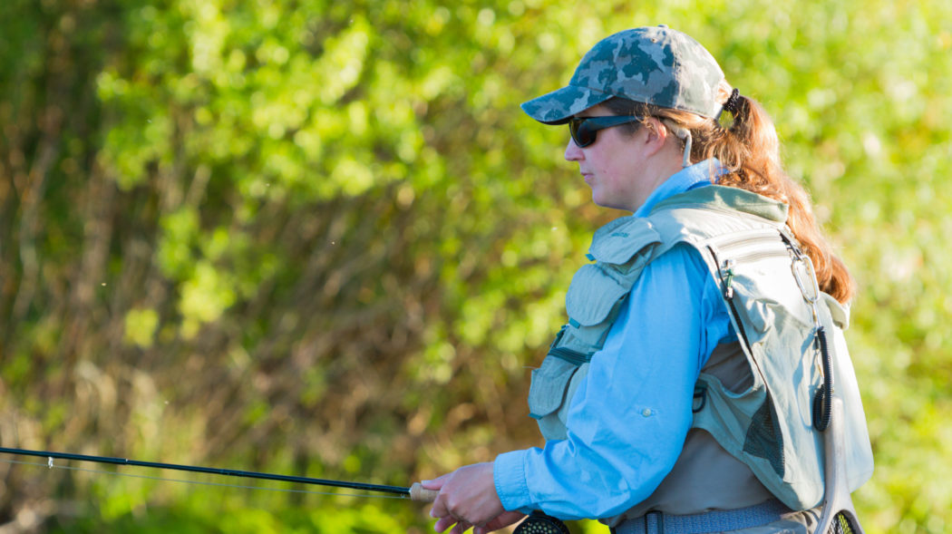 Woman Fly Fishing for trout In A Yellowstone Creek. She is wearing waders, a fly fishing vest and a camouflage hat.