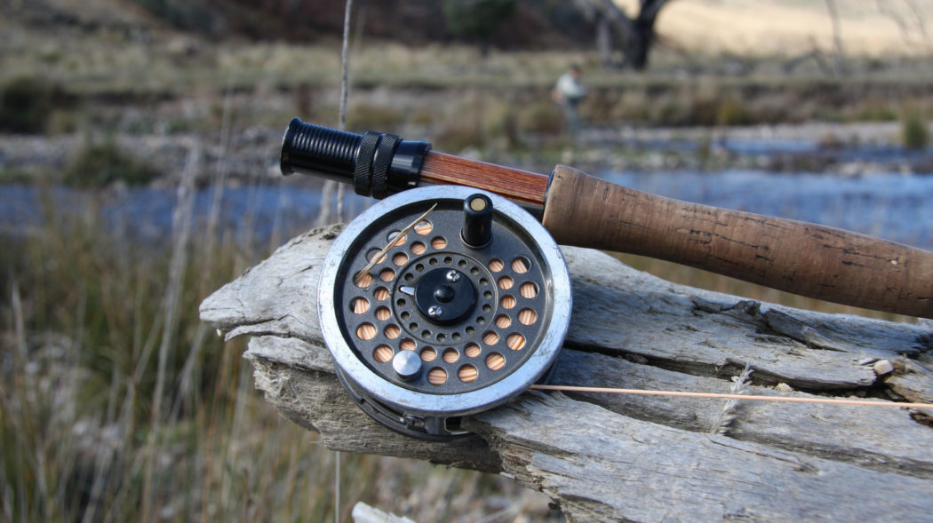 A picture of a fly fishing reel on a fly rod, resting on a log near a trout stream. The blurred form of an angler can be seen in the background