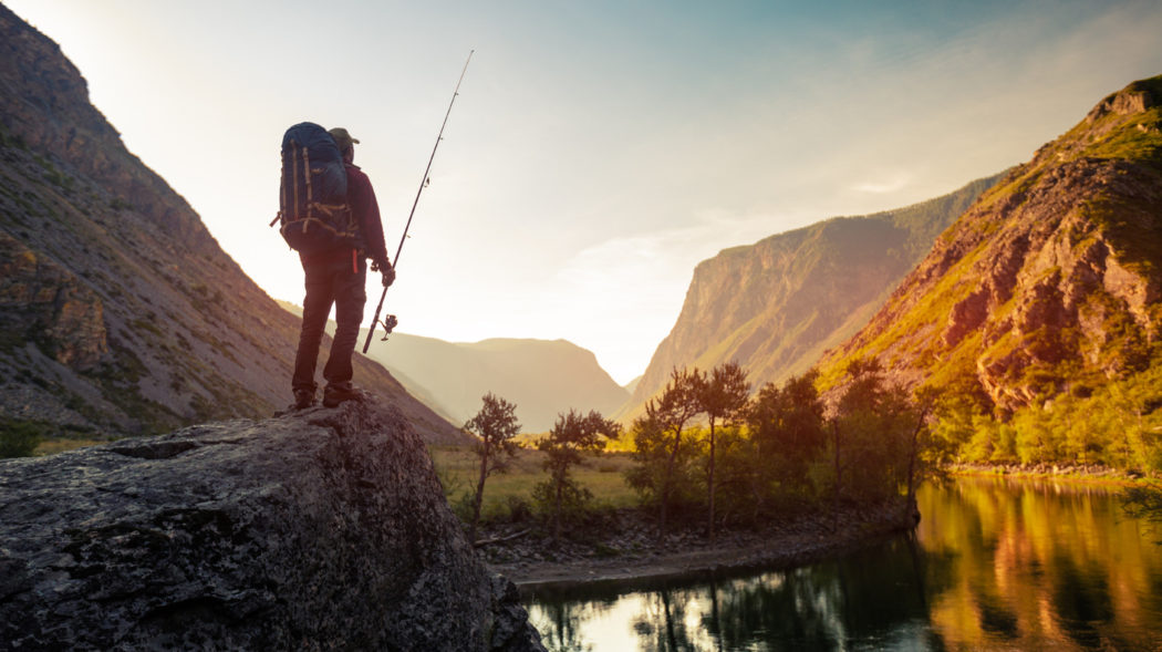Hiker stands on the rock with fishing backpack and fishing rod and enjoys sunrise river view